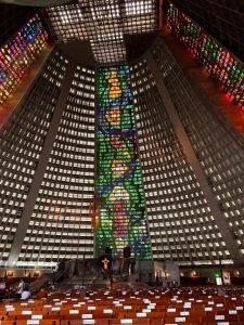 The image showcases the interior of a large, modern church or cathedral featuring impressive stained glass windows. The walls are lined with geometric shapes and large panes of colored glass that create vibrant patterns of blue, green, red, and yellow light. In the foreground, rows of wooden pews are arranged neatly, some with labels attached. At the altar, there is a simple wooden cross and a partially covered statue, against a backdrop of a strikingly designed wall. The ceiling is high, with a grid-like structure that complements the contemporary architectural style. Overall, the ambiance is serene and contemplative.