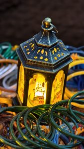 A black and gold lantern with Arabic calligraphy glows warmly, sitting on colorful glass bangles against a dark blurred background.