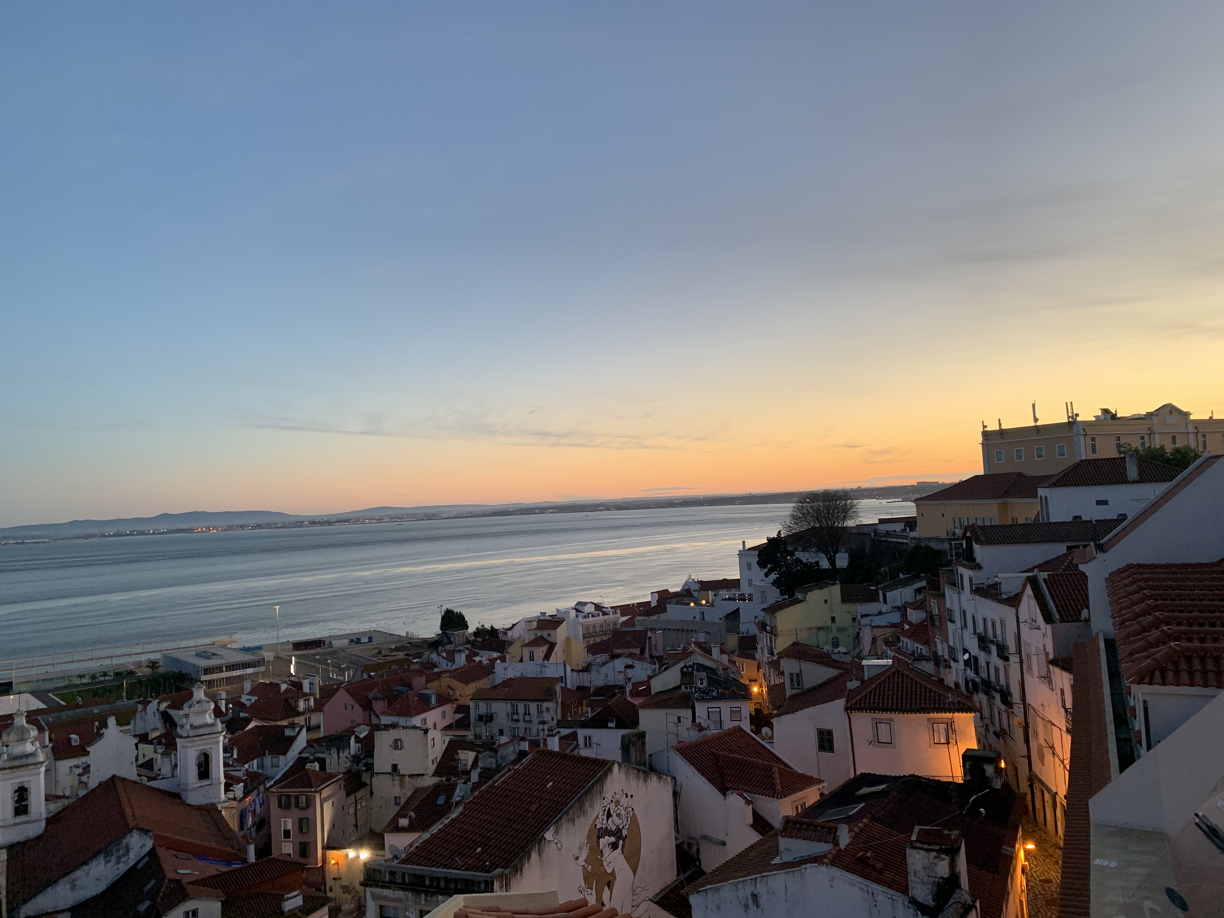 Alfama neighborhood seen from above, with white houses and terracotta roofs, next to the ocean and the sunset. Lisbon, Portugal.