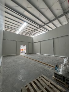 A spacious, empty interior of a warehouse or storage facility with high ceilings and gray walls. Natural light streams in through large windows in the roof, illuminating the concrete floor. At the far end, a double door opens to an area outside, suggesting an operational environment. A wooden pallet is positioned on the right side, and to the right of it, there are bags or materials stacked on a gray block.