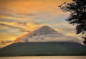 A picturesque view of a mountain with a cloud-covered peak, set against a vibrant sunset sky that transitions from orange to blue. The foreground includes a body of water reflecting the colors of the sky, while a tree on the right adds a natural element to the scene. The overall atmosphere is tranquil and scenic. - Acatenango Volcano