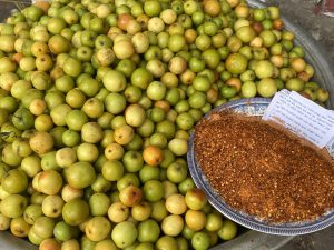A pile of green and orange fruits beside a plate of red chili powder and papers.