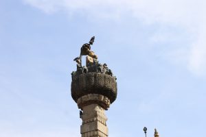 A tall, intricately carved pillar topped with a decorative sculpture featuring various animal figures and mythical beings against a clear blue sky.
