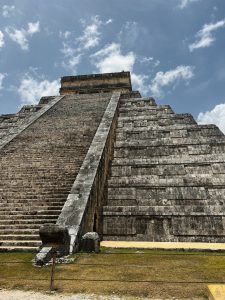Chichen Itza Pyramid, one of the modern wonders of the world. The pyramid is made of gray stone and features distinct layers. In the foreground, there is a green lawn and a metal barrier.
