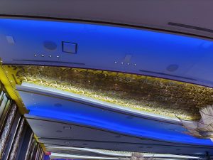 Blue-lit ceiling with a speaker, rectangular vent, four spotlights, a linear vent, and a gold decorative panel with swirls.