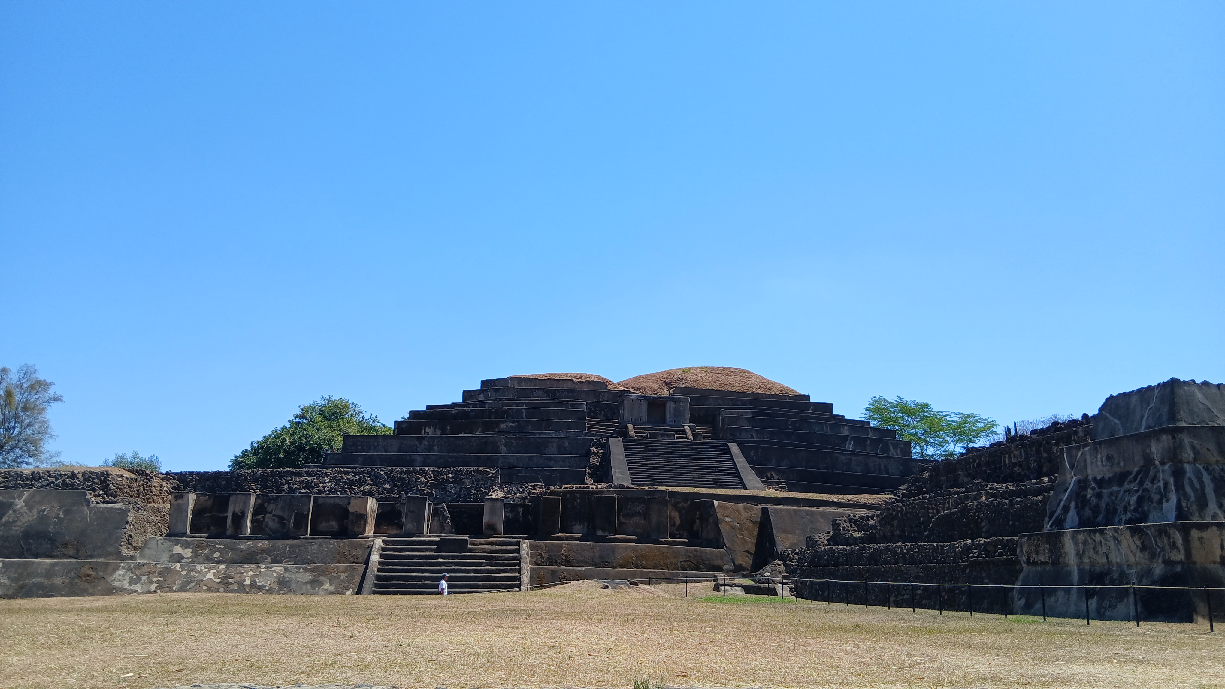 A large, ancient pyramid structure is set against a clear blue sky. The pyramid features multiple tiers with staircases leading to the top. Surrounding the pyramid are stone walls and grassy areas, with a few trees in the background. In the foreground, a small figure is visible, adding a sense of scale to the scene.