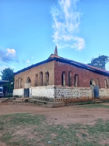 An old plastered church building with narrow arched windows, a rusted roof, and a dirt yard under a blue sky.