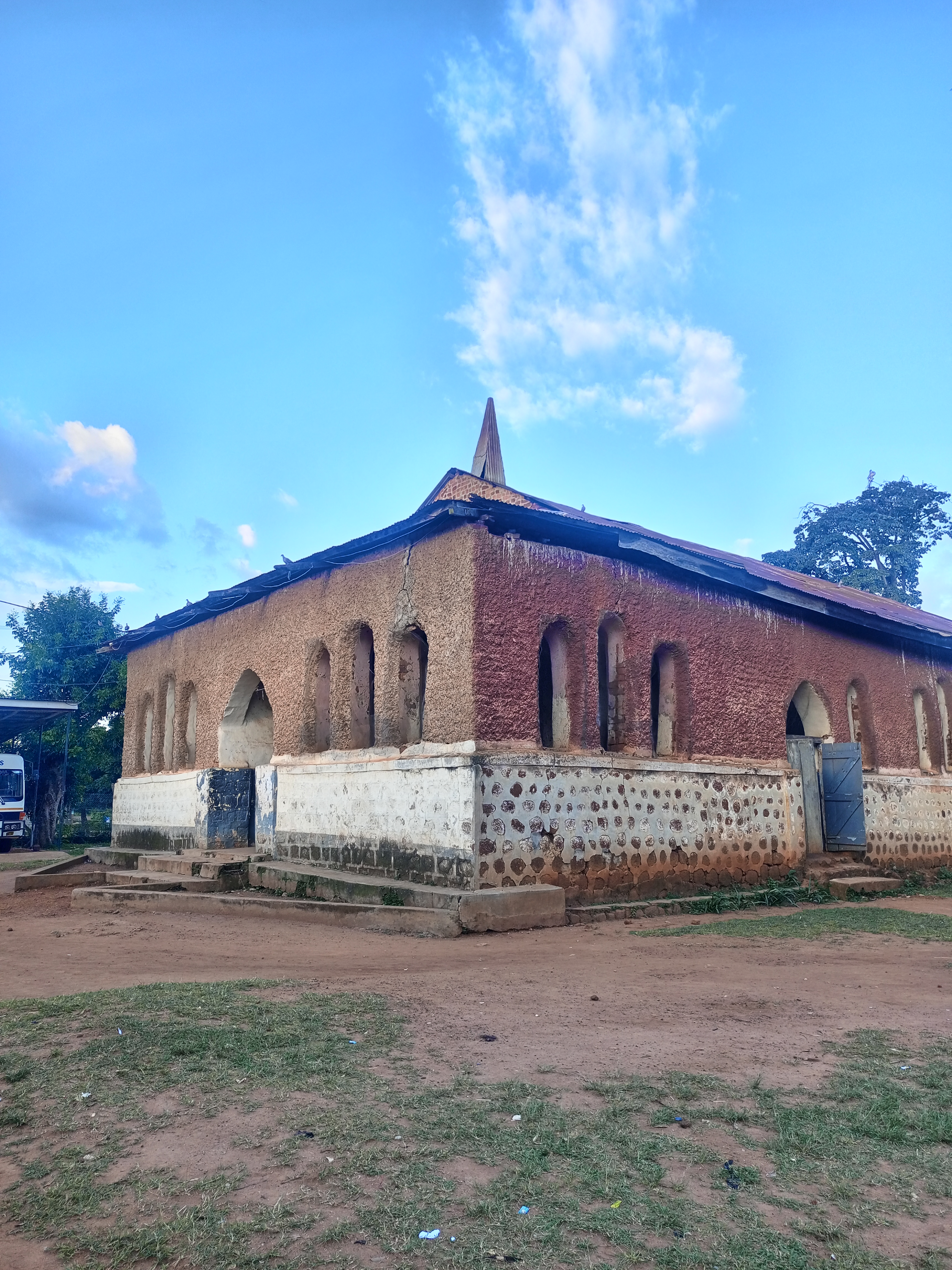 An old plastered church building with narrow arched windows, a rusted roof, and a dirt yard under a blue sky.