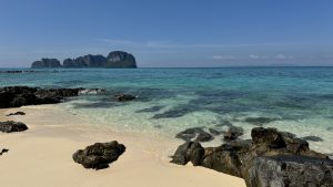 A peaceful beach scene shows clear shallow water washing over dark rocks on the shore. The sand is very pale and clean. In the background, a group of rocky islands sits in the middle of the turquoise ocean. The sky is bright blue and completely clear of clouds.