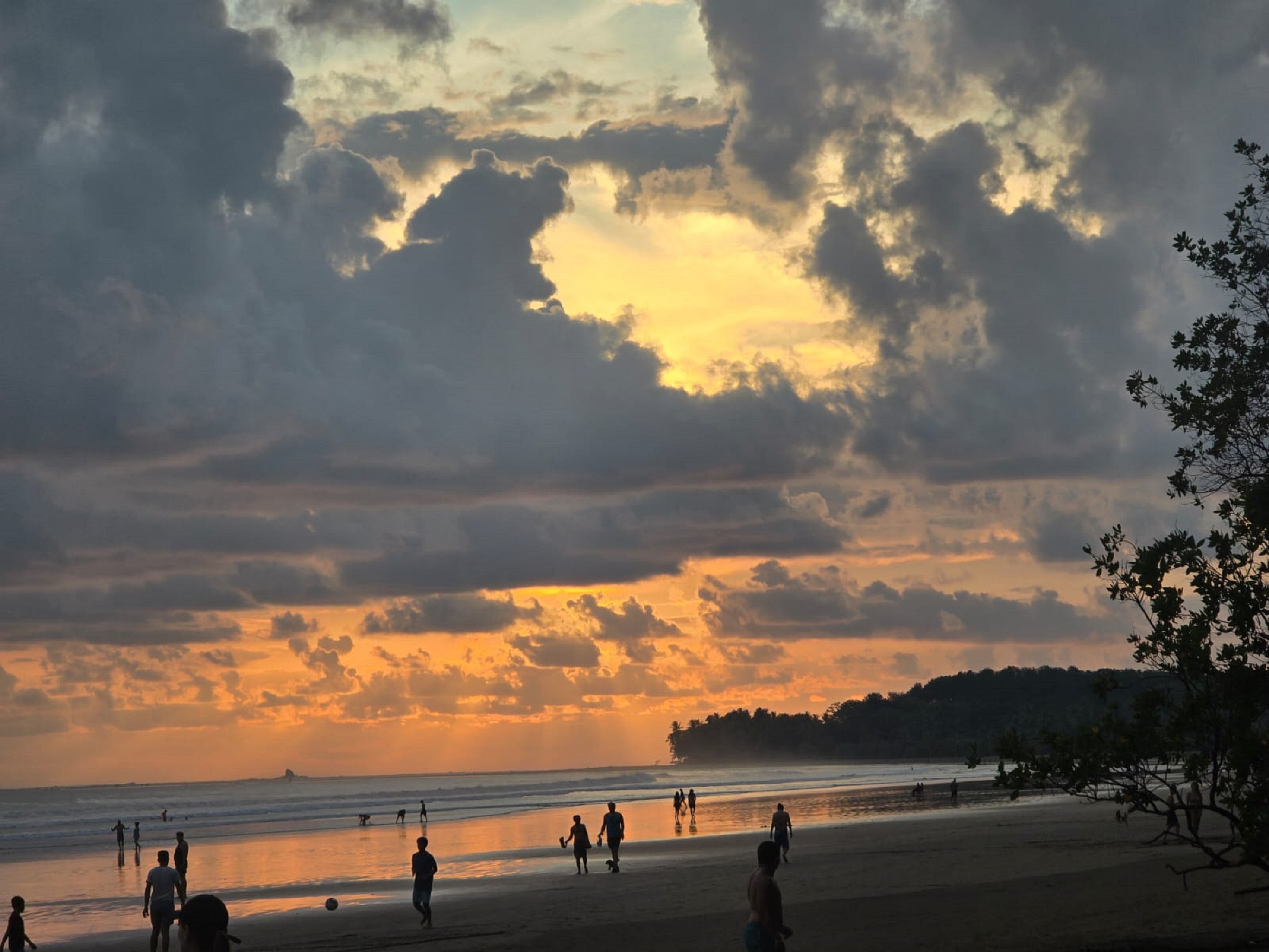 Beach at sunset with people walking on the wet sand under a dramatic sky filled with clouds and golden light.
