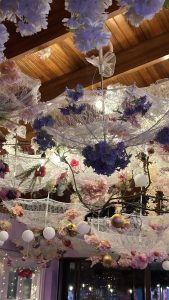 The image features a whimsical decor scene with white lace umbrellas suspended from a wooden ceiling, adorned with various artificial flowers in shades of pink and purple. The umbrellas are interspersed with small, decorative orbs, adding a festive touch. Soft lighting enhances the ambiance, creating a romantic and enchanting atmosphere.