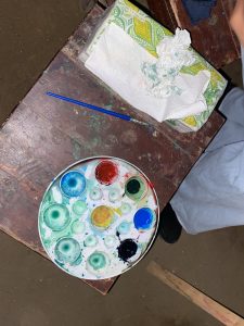 A colorful paint palette with mixed hues, a blue brush, and crumpled tissues on a wooden table suggests a vibrant, creative art session.
