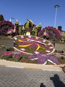 A large outdoor floral display shaped like a clock, covered in vibrant pink flowers with oversized numbers and clock hands. Surrounding it are landscaped gardens, colorful blooms, and decorative structures resembling castles and peacocks, set under a clear blue sky.
