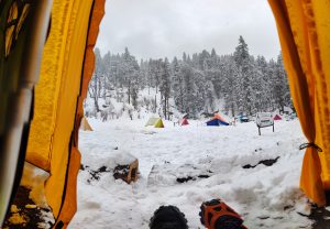 View from inside a tent opening onto a snowy campsite, with colorful tents scattered across a snow-covered clearing and a forest of snow-laden pine trees in the background under an overcast sky.