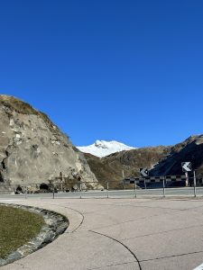 A winding road curves around a rocky landscape, with a clear blue sky overhead. In the background, a snow-capped mountain peak stands majestically against the horizon. The foreground features grassy patches and a stone-lined path, while road signs indicate a turn ahead.