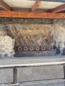 A section of an ancient wall painting featuring a stylized animal figure, likely a feline, surrounded by red and white abstract patterns. The artwork is set against a rough stone wall and is partially obscured by weathered plaster. Above, wooden beams of a roof are visible, indicating the painting is located indoors or under a protective structure.