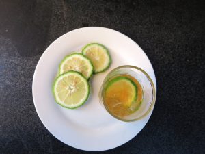 A white plate holds three slices of green citrus fruit, with a glass of light-brown drink and another slice beside it on a dark countertop.