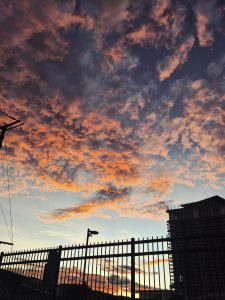 A vibrant sunset sky with orange, pink, and purple clouds over a city scene, with a dark fence, streetlights, and a building silhouette in the foreground.