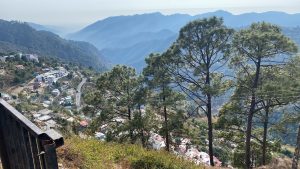 A breathtaking mountain landscape with layered blue hills fading into the distance, a winding road through a peaceful hillside town, colorful houses, tall pine trees in the foreground, soft sunlight over the valley, slight haze, cinematic depth, ultra-realistic, 4K, serene wide-angle view
