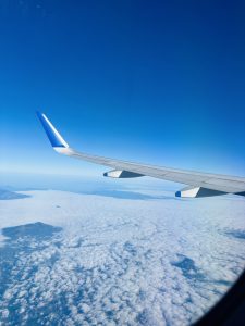 View from an airplane window showing a wing over white clouds and distant mountains under a clear blue sky.