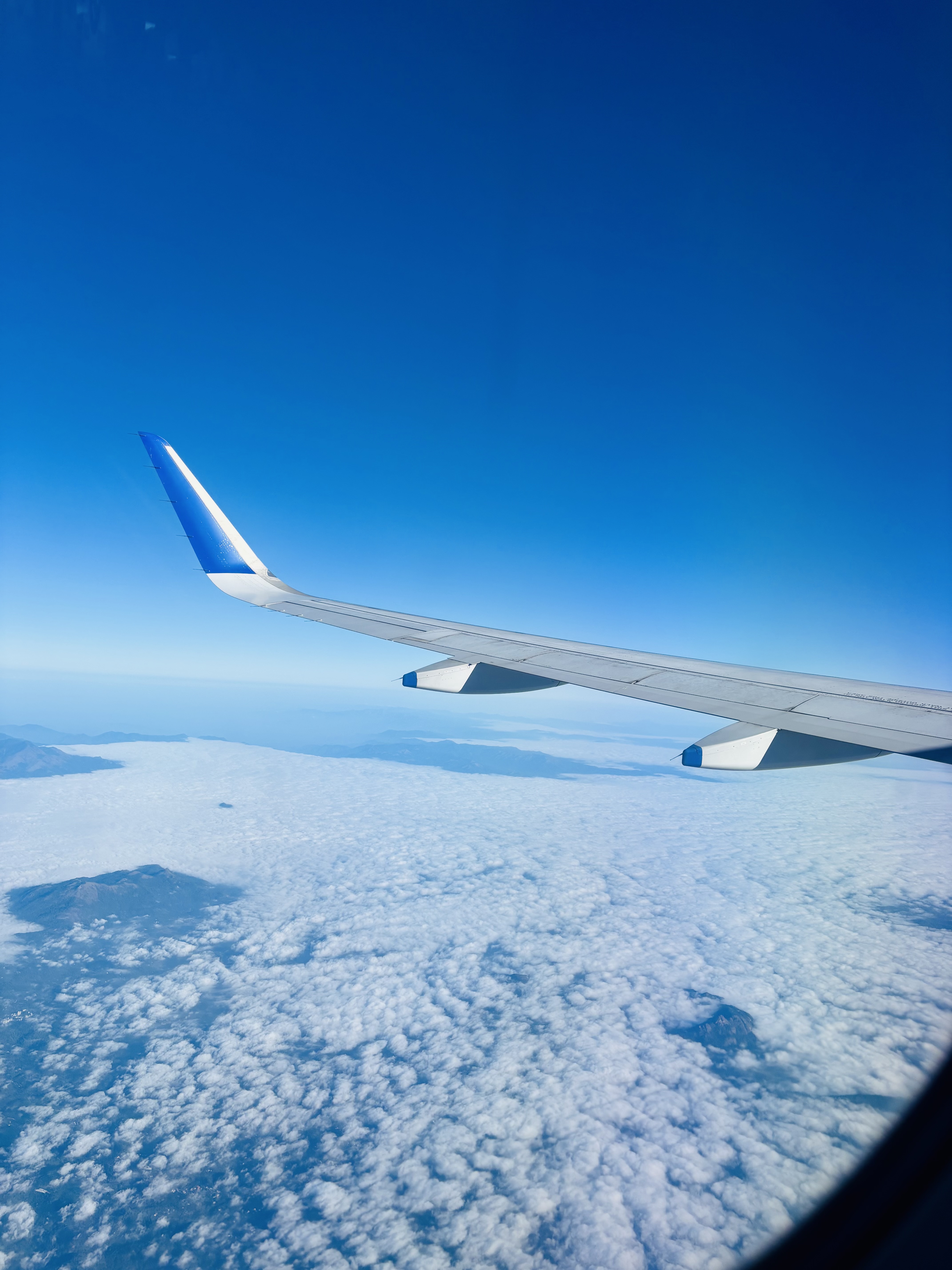 View from an airplane window showing a wing over white clouds and distant mountains under a clear blue sky.