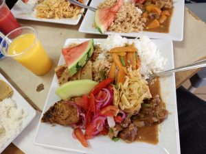 A white plate holds rice, pilau, beef stew, fried chicken, cabbage, topped with melon slices, with orange juice on the side.