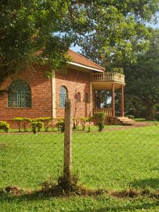 A red-brick house featuring a balcony and decorative columns sits nestled within lush, well-maintained greenery. Tall trees and a chain-link fence frame the serene property, highlighting its large windows and classic architectural details.
