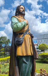 A large, colorful statue of a man with long hair and a beard, dressed in a brown robe and a green shirt, stands with his right hand over his heart. He has a black bag slung across his body and a backdrop of a bright blue sky filled with clouds. Lush greenery and shrubs surround the statue, with some buildings visible in the distance.