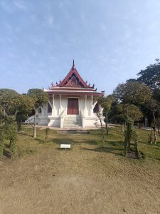 White monastery with an ornate red roof, centered on a grassy hill under a blue sky.
