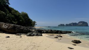 A wide view of a tropical coastline shows a white sand beach with many dark rocks near the water. On the left, there is a dense green forest. The ocean is clear turquoise and very calm. In the far distance, a large rocky island with green plants sits under a clear blue sky.