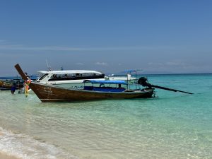 Several boats rest in shallow, clear light green water near a white sand beach. A wooden longtail boat with a blue roof sits in front, with a larger white speedboat behind it.