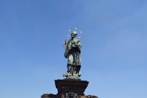 A bronze statue of a saint with a golden star halo and a palm leaf, set against a clear blue sky in prague.
