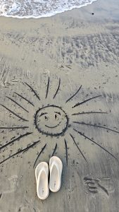 White flip-flops on a sandy beach beside a smiling sun drawing, with gentle ocean waves in the background.
