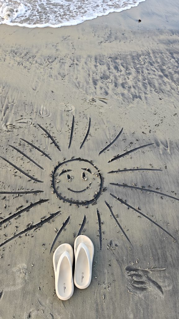 White flip-flops on a sandy beach beside a smiling sun drawing, with gentle ocean waves in the background.