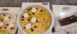 Creamy yellow desserts/ cakes decorated with white cream swirls, blueberries, and seeds, along with a chocolate cake placed on the side.
