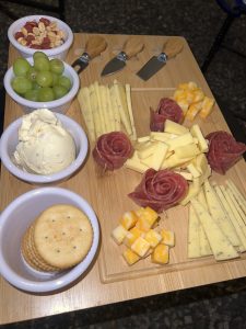 A wooden serving board features an assortment of snacks including bowls of green grapes, mixed nuts, and a creamy spread. There are arranged slices of yellow cheese, along with rolled slices of salami. Round crackers are neatly placed on the board.