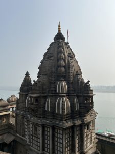 A Maheshwar stone temple with ornate carvings and a tall spire stands on the riverbank, with water and hazy sky in the background.