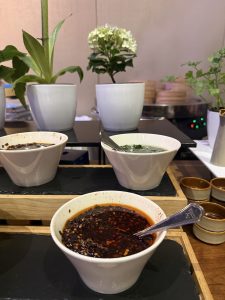 A wooden serving tray features three small white bowls containing different sauces, with one bowl filled with a dark, spicy sauce garnished with herbs