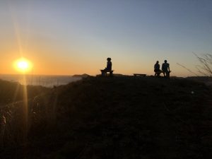 Mountain summit with human silhouettes watching the sunset over the beach.