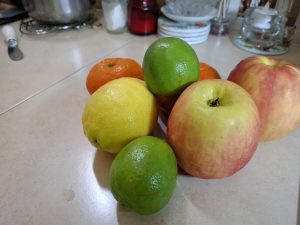 
A collection of various fruits, including two red and yellow apples, a yellow lemon, two green limes, one orange, and a tangerine, arranged on a neutral kitchen countertop. 