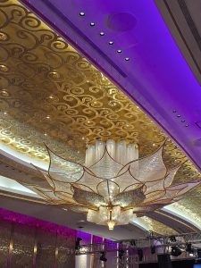 An ornate flower-shaped chandelier with intricate gold detailing and sparkling crystals hangs from a richly decorated ceiling with swirling gold patterns, illuminated by soft purple lighting and complemented by modern fixtures, creating a luxurious and sophisticated ambiance.