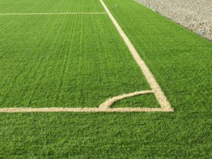 White corner line on bright green artificial turf with visible synthetic grass texture, bordered by grey gravel and stones.