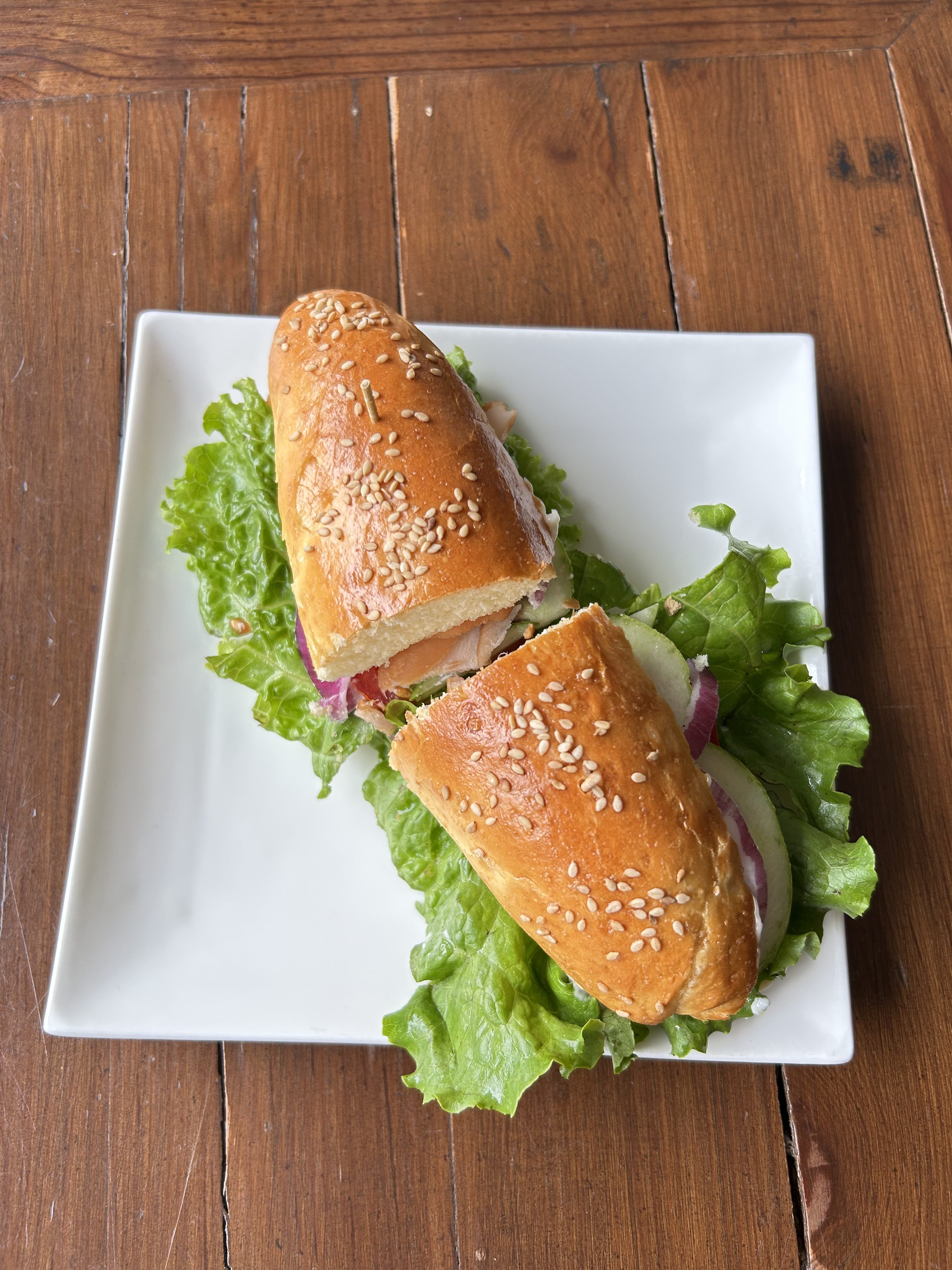 A close-up view of a freshly made sandwich, served on a white square plate. The sandwich features a golden-brown, sesame seed-topped bun, split open to reveal layers of ingredients inside.