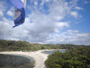 A scenic view of a beach surrounded by lush greenery and a clear sky with scattered clouds. In the foreground, there is a flag waving, and in the distance, people are seen enjoying the beach. Bahía Ballena.