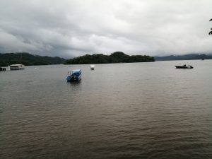 A calm body of water stretches across the scene under a heavy, overcast sky. Small boats float quietly on the surface, with lush green hills and small islands in the background partially covered by mist.