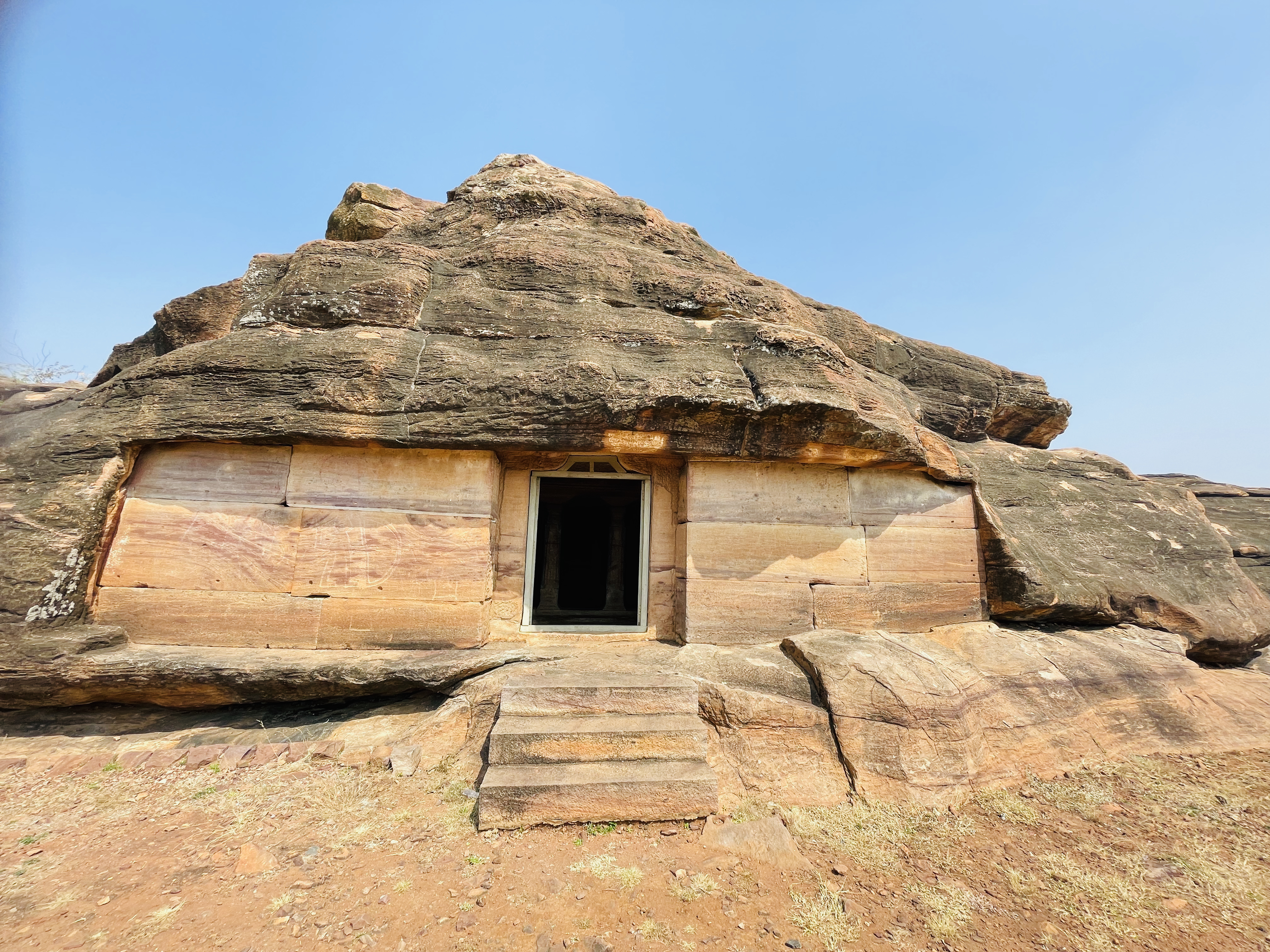 A rock-cut structure with a doorway built into natural stone. The earthy tones and rugged shape give a unique, ancient look. Captured at Aihole, Bagalkot, Karnataka.