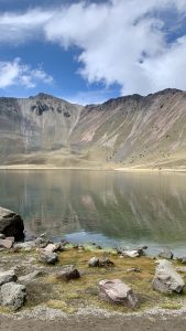 The still surface of Laguna del Sol reflects the steep crater walls of Nevado de Toluca, with rocky shores, sparse alpine vegetation, and tiny figures in the distance showing scale in Estado de México.
