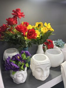 A decorative arrangement of artificial flowers in vases, including red, yellow, and purple blooms, with white ceramic face-shaped vases on a shelf.