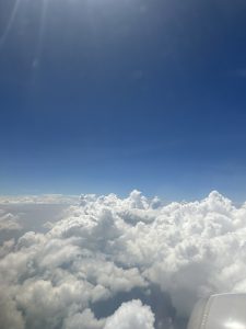 A view from an airplane window showing a vast sky filled with fluffy white clouds under bright blue skies, with the sun shining brightly in the upper left corner.

