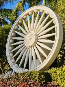 A large Ashoka Chakra wheel sculpture surrounded by greenery at Global Vipassana Pagoda, Gorai Village, Mumbai. The bright light and strong shapes make it visually striking.
